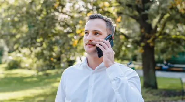 Ein Mann befindet sich in einem sonnendurchleuchteten, grünen Park und blickt in die Ferne während er ein Telefonat mit einem Berater führt.